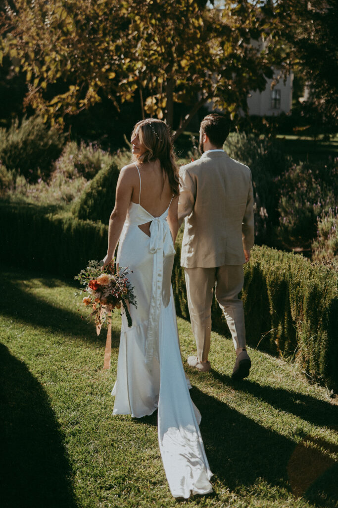 A bride in a white dress holding a bouquet walks on grass beside a groom in a light suit, with sunlight and greenery around them, capturing the magic of a fall park wedding.