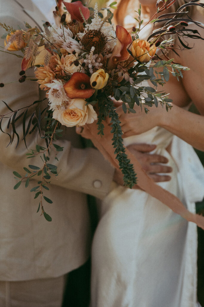 A person in a light suit embraces another in a white dress holding a bouquet of orange, yellow, and white flowers with greenery, capturing the warmth of a Fall Park wedding.