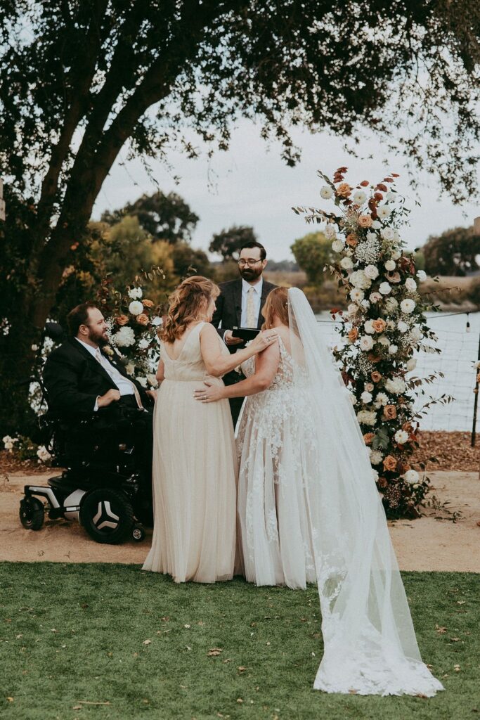 A wedding ceremony by a lake with two brides in white dresses and a person in a suit officiating. One guest, seated in a wheelchair, is part of the event. Floral arch in the background.