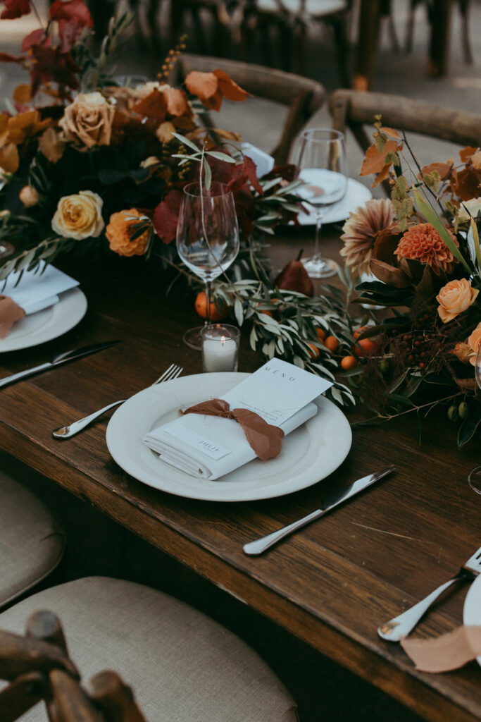 Elegant table setting with a white plate, menu card, napkin, brown leaf, and an autumn floral arrangement on a wooden table—perfect for a cozy Fall Park Winters Wedding celebration.