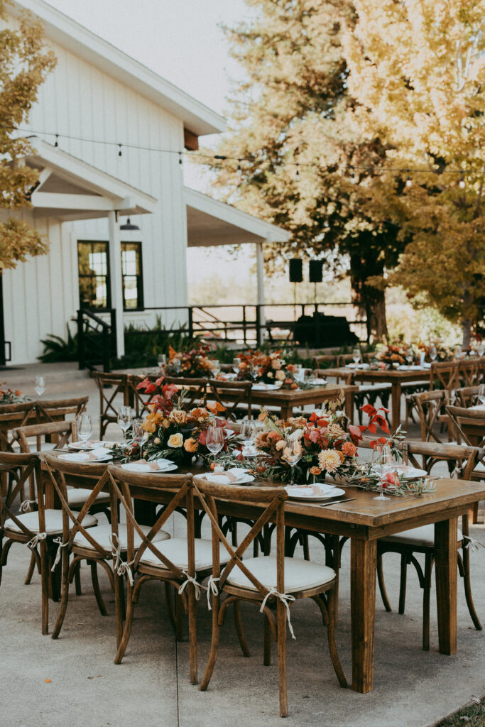 Outdoor dining area with wooden tables and chairs, decorated with flower centerpieces, set beside a white building surrounded by trees—an inviting setting perfect for a Fall Park wedding or cozy Winters celebration.