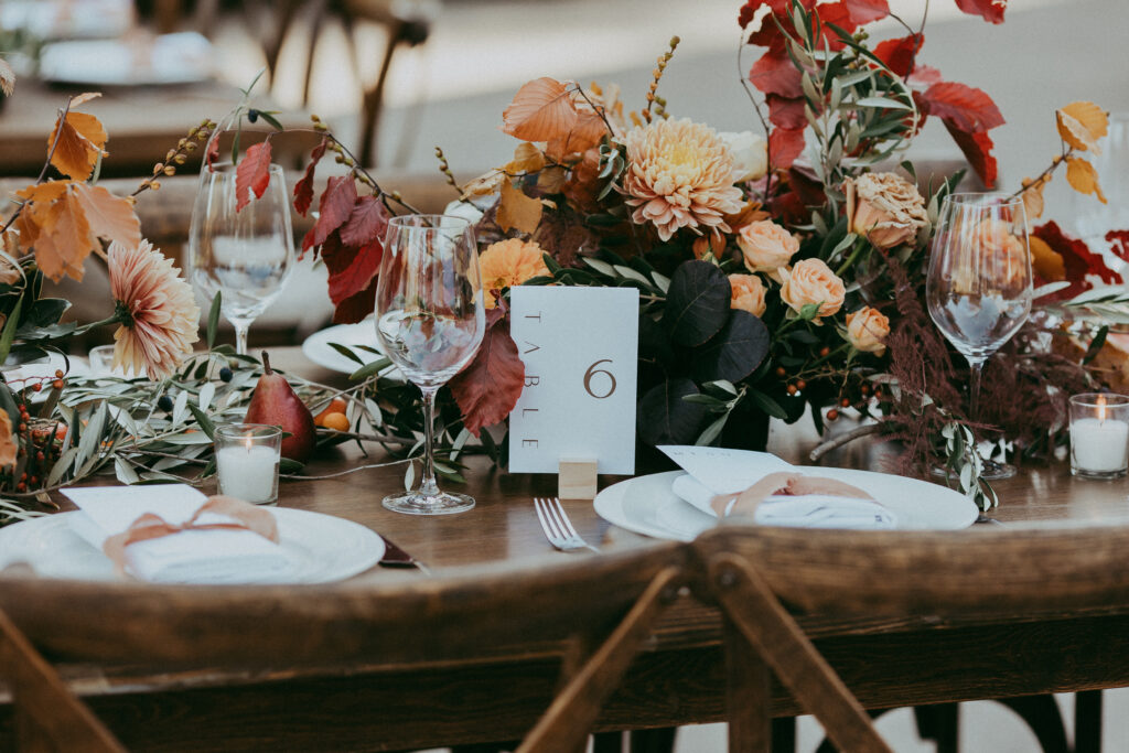 Table set for an event with a fall-themed floral centerpiece, wine glasses, plates, candles, and a card reading “Table 6,” perfect for a cozy Fall Park Winters Wedding.