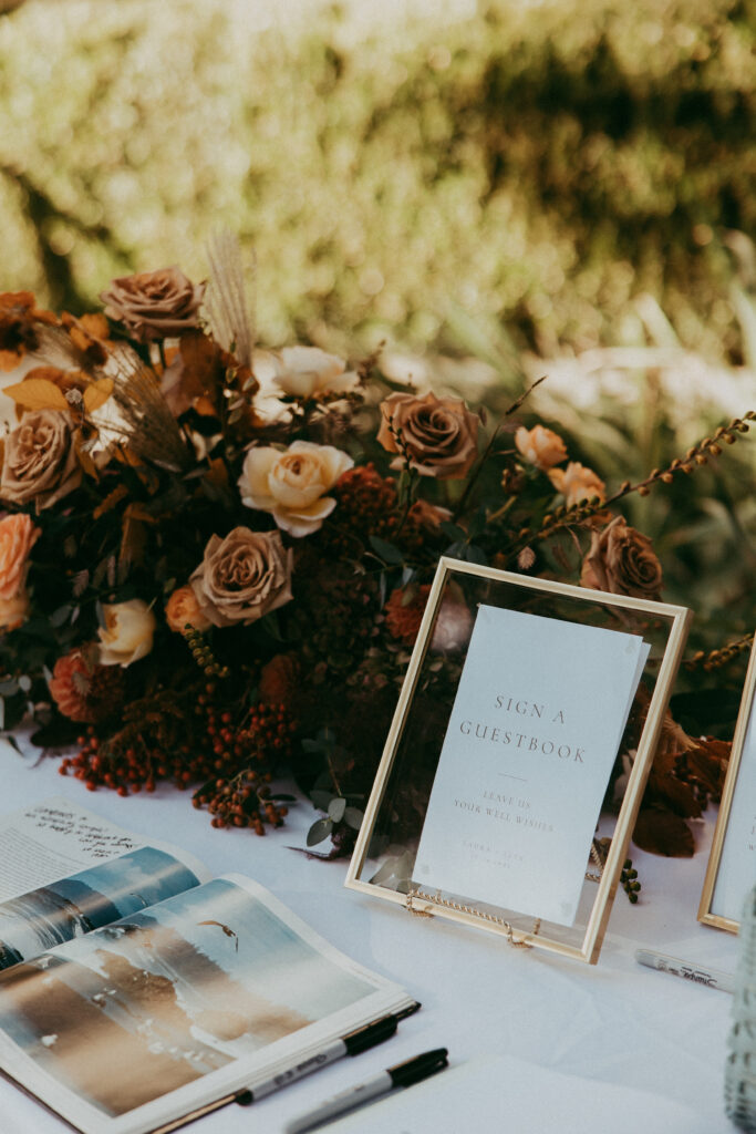 A guestbook and pens on a table with a sign reading “Sign a Guestbook,” next to a floral arrangement with brown and beige roses, perfect for a cozy Fall Park Winters wedding.
