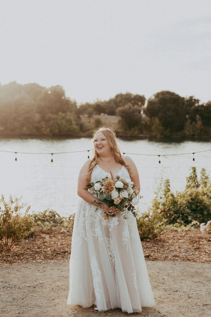A bride in a white dress stands outdoors, smiling while holding a bouquet. Behind her, a body of water and trees under the sunlight are visible.