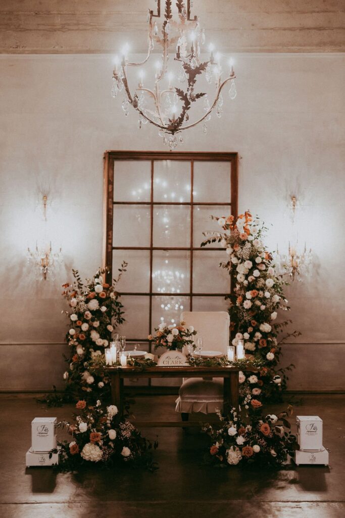 A wedding table is elegantly decorated with floral arrangements, candles, and a chandelier overhead. A backdrop of wooden window panes adds a rustic touch.