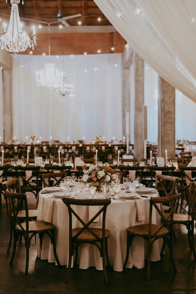 Elegant wedding reception with a round table set for guests, featuring floral centerpieces and lit candles. Chandeliers and draped fabric create a soft, romantic ambiance.