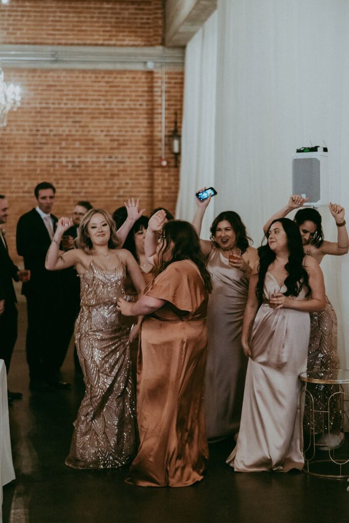 A group of people in formal attire are dancing and smiling at an indoor event with brick walls in the background.