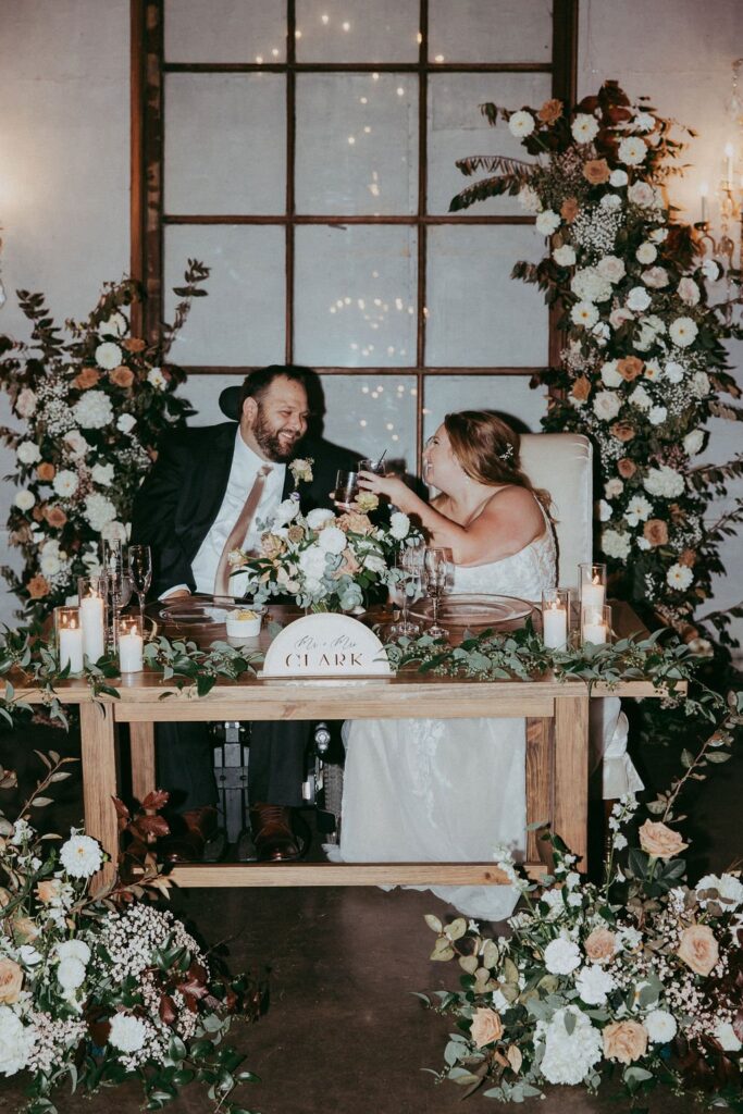 A couple in wedding attire clinks glasses while seated at a table adorned with flowers and candles. A sign reads "Mr. & Mrs. Clark.