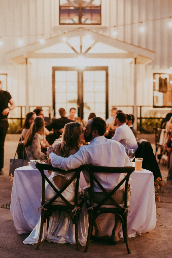 A couple sits closely at a table, backs to the camera, during an outdoor evening event in a fall park, with other guests in the background and string lights overhead.