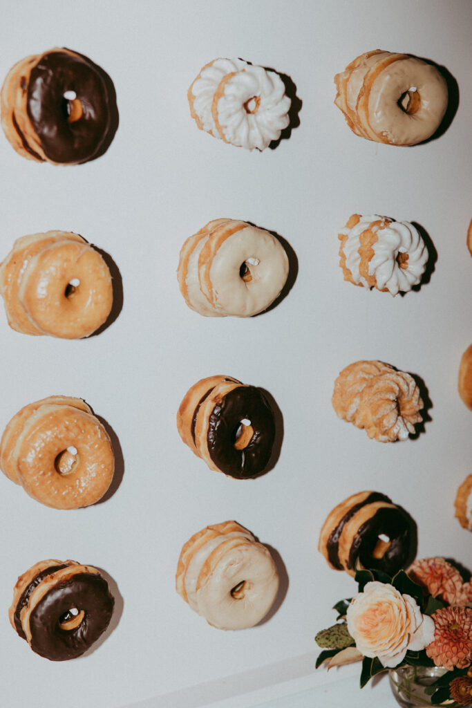 Assorted donuts, including glazed and chocolate, arranged in even rows on a white wall; a small floral arrangement is visible in the lower right corner, creating a charming display perfect for a Fall Park Winters wedding.