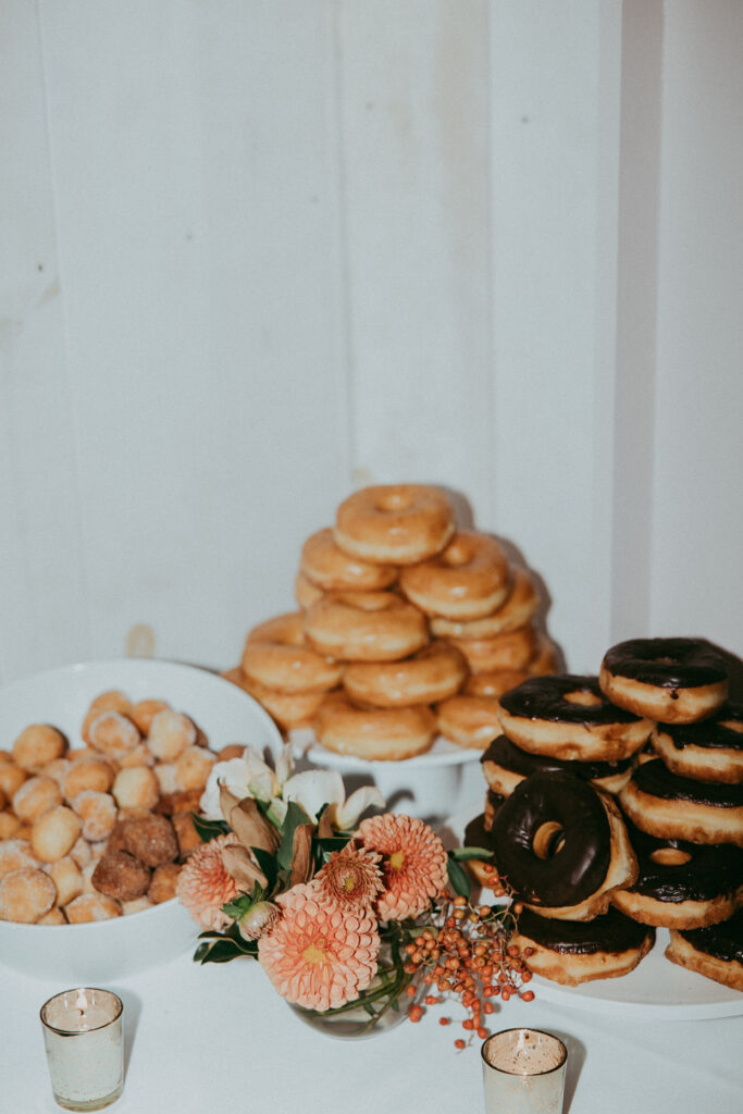 Three stacks of assorted donuts and donut holes on a table, with a small floral arrangement and two small votive candles in the foreground, create a cozy scene perfect for a Fall Park Winters wedding.