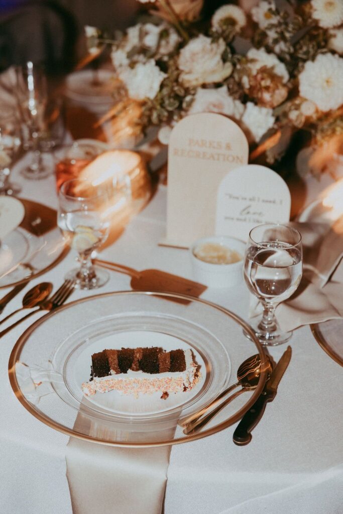A slice of chocolate cake on a glass plate sits on an elegant table with a floral centerpiece, glassware, utensils, and a "Parks & Recreation" card.