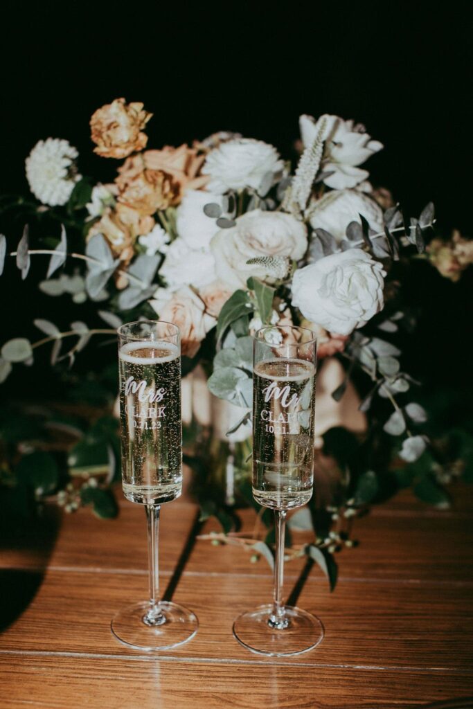 Two champagne flutes engraved with numbers and text are placed in front of a floral bouquet with white and peach roses on a wooden surface.