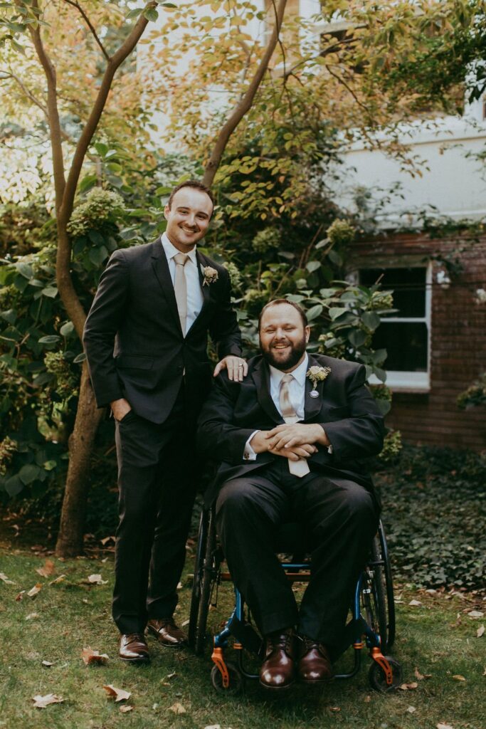 Two men in suits, one sitting in a wheelchair and the other standing beside him, pose for a photo in a garden with trees and greenery.