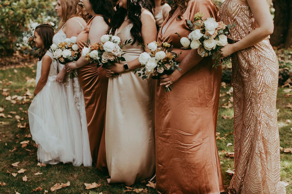 Group of women and a young girl in formal dresses holding bouquets, standing outdoors on grassy ground with fallen leaves.