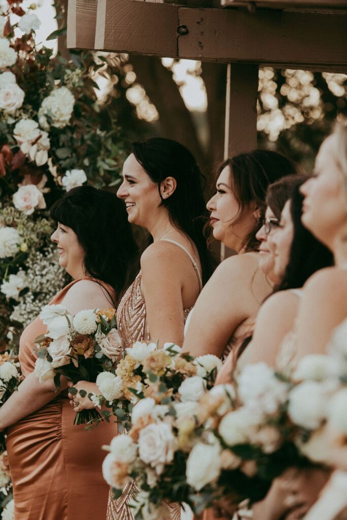 A group of women in formal dresses hold floral bouquets, standing outdoors near a wooden structure adorned with more flowers.