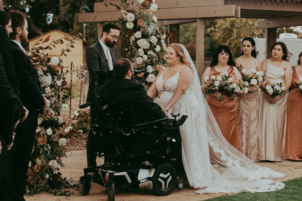 Bride and groom exchange vows under a floral arch, surrounded by bridesmaids holding bouquets. The groom is in a wheelchair.