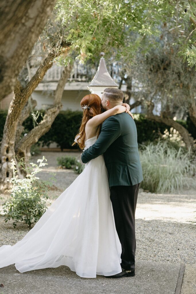 A bride and groom embrace outdoors, surrounded by trees and greenery.