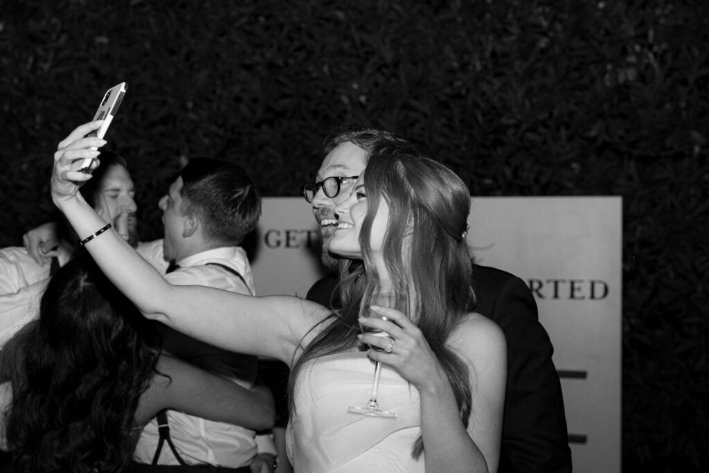 A couple takes a selfie at an event, with one holding a phone while the other has a glass. Other people embrace in the background.