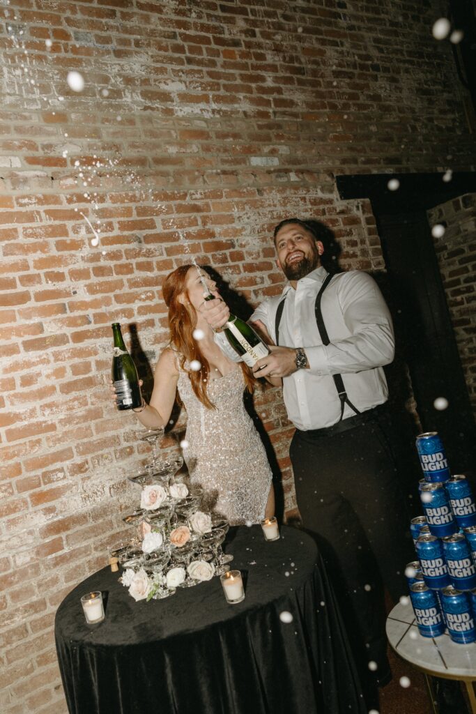 A couple happily sprays champagne in front of a brick wall, standing next to a table with flowers, candles, and a stack of beer cans.