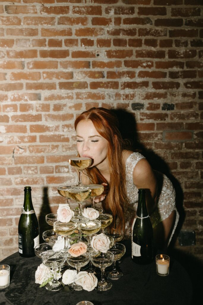 A woman in a dress leans in to drink from a top champagne glass of a pyramid stack. Bottles and candles are on a nearby table against a brick wall.