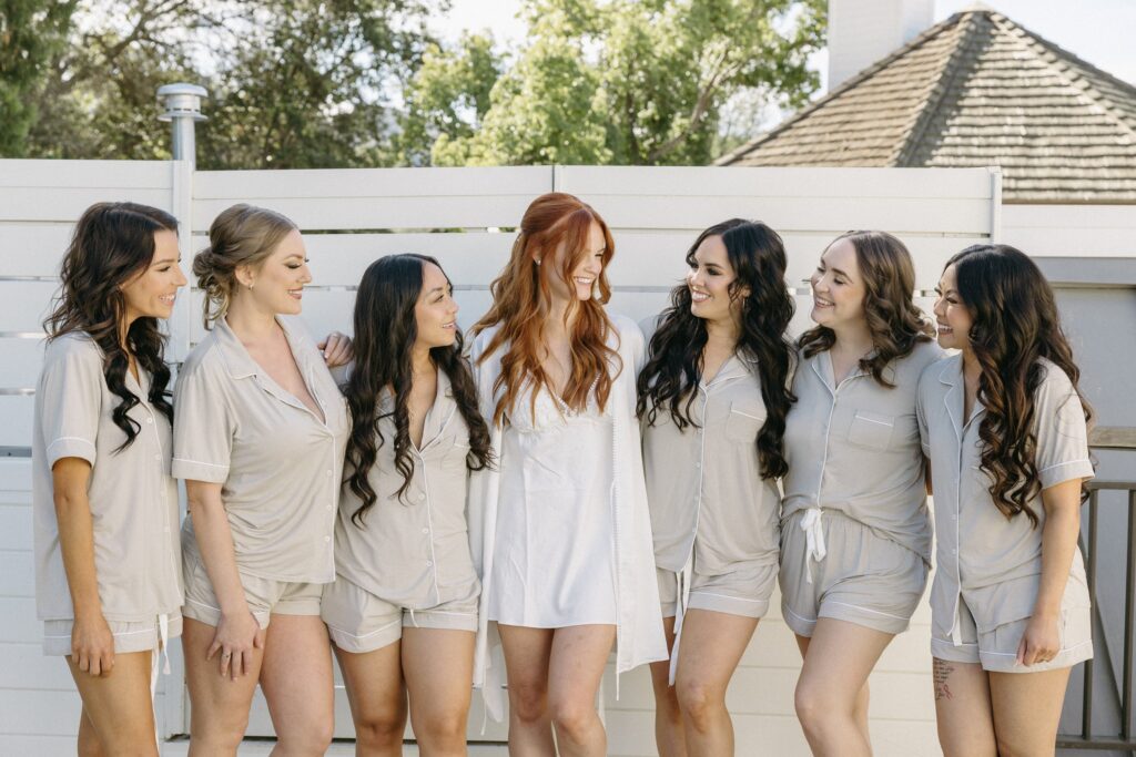 Seven women in matching loungewear stand together outdoors, with one woman in a white robe at the center, smiling and facing each other.