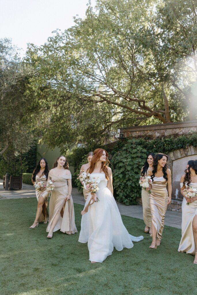 A bride in a white dress walks with bridesmaids in gold dresses on a grassy area, surrounded by trees and greenery.