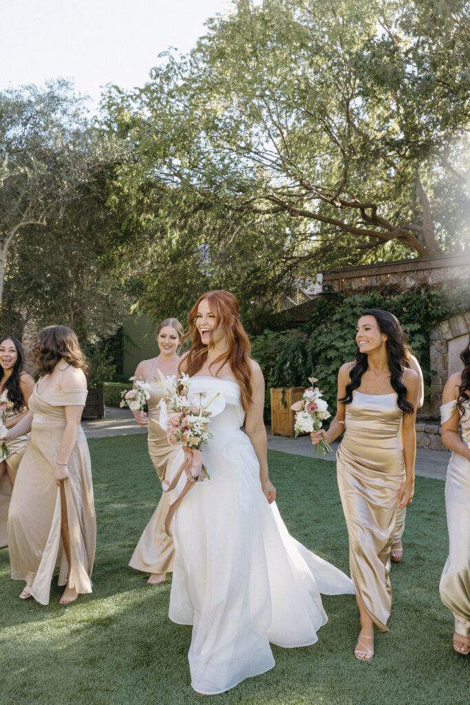 A bride in a white dress walks with bridesmaids in gold dresses on a lawn, surrounded by trees.