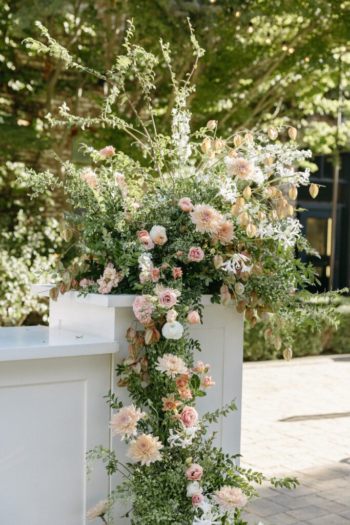 Floral arrangement with pink and white flowers and green foliage on a white stand outdoors.