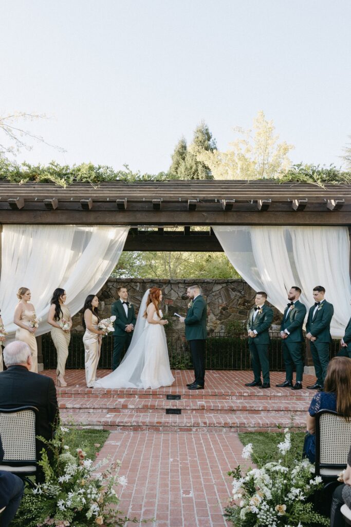 A wedding ceremony outdoors with a couple exchanging vows under a pergola. Bridesmaids in gold dresses and groomsmen in green suits stand beside them. Guests are seated in the foreground.