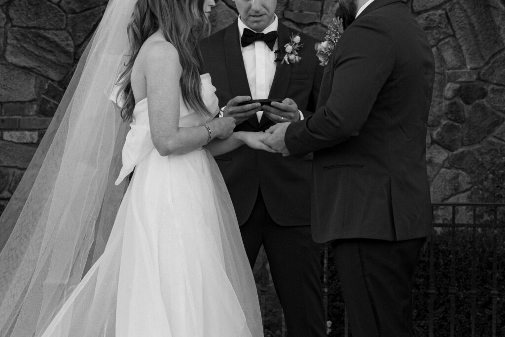 A bride and groom hold hands during their wedding ceremony, with an officiant reading from a device. They stand in front of a stone wall.