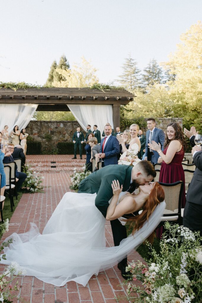 A groom kisses his bride, dipping her backward, in an outdoor wedding ceremony. Bridesmaids, groomsmen, and guests stand around, smiling and clapping.