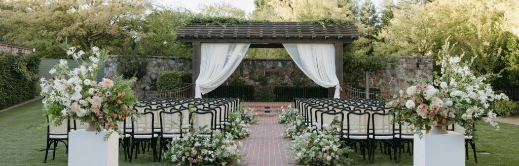Outdoor wedding ceremony setup with rows of black chairs facing a pergola draped in white fabric, surrounded by greenery and floral arrangements on a brick pathway.