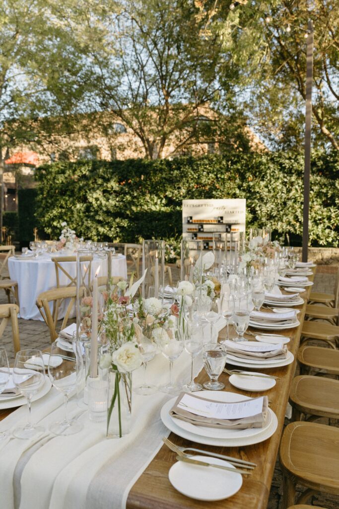 Elegant outdoor dining setup with a long wooden table, white floral centerpieces, candles, and neatly arranged tableware. Surrounded by greenery and wooden chairs.