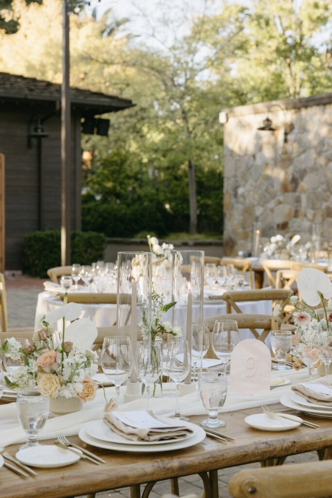 Outdoor wedding reception setup with rustic wooden tables, elegant white and beige floral arrangements, white plates, and tall glass candle holders against a stone wall backdrop.