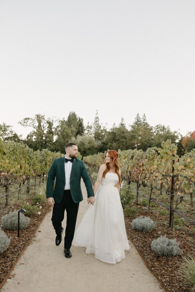 A couple in formal attire walks hand in hand along a path in a vineyard.