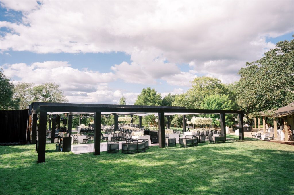 Outdoor event setup with black wooden canopy, chairs, and tables on a grassy area, surrounded by trees under a partly cloudy sky.