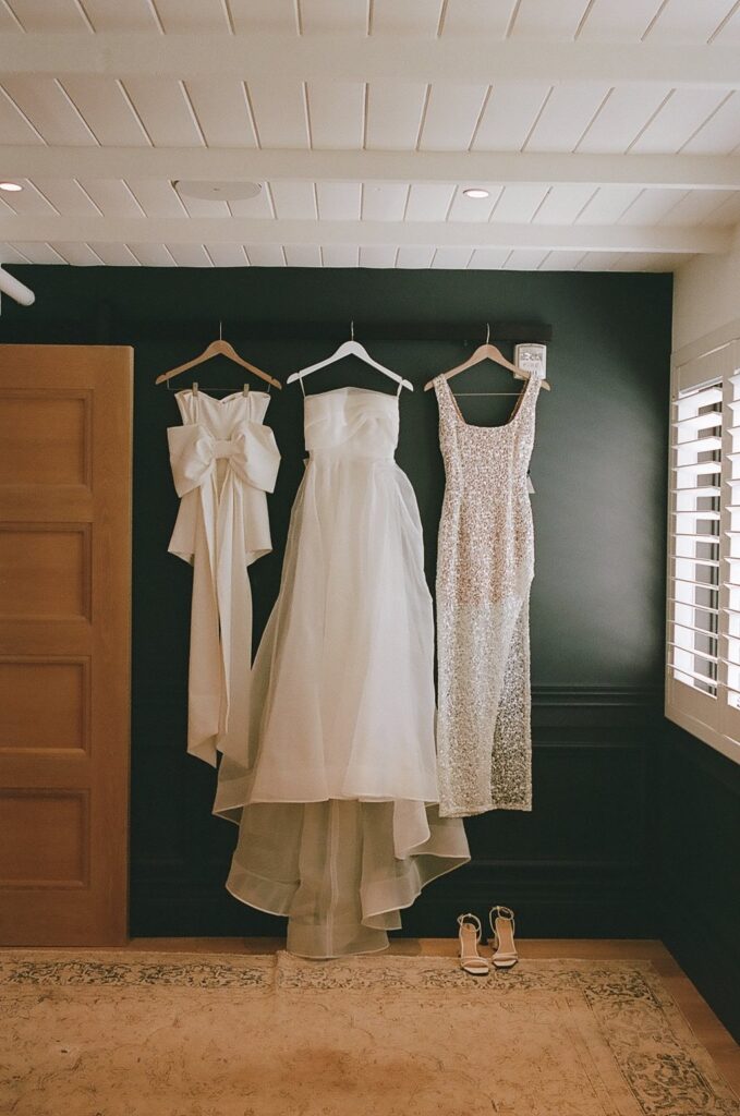Three dresses, including a strapless gown and a lace dress, hang on a dark wall with a pair of sandals below.
