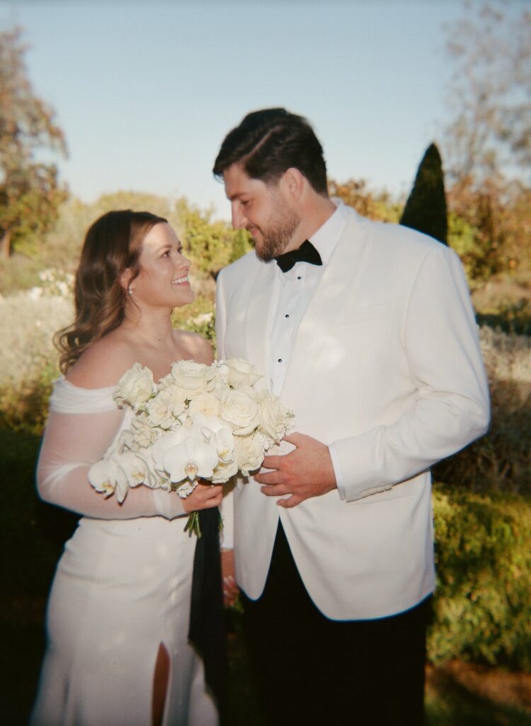 A couple dressed in formal attire stands outdoors, smiling at each other. The woman holds a bouquet of white flowers.