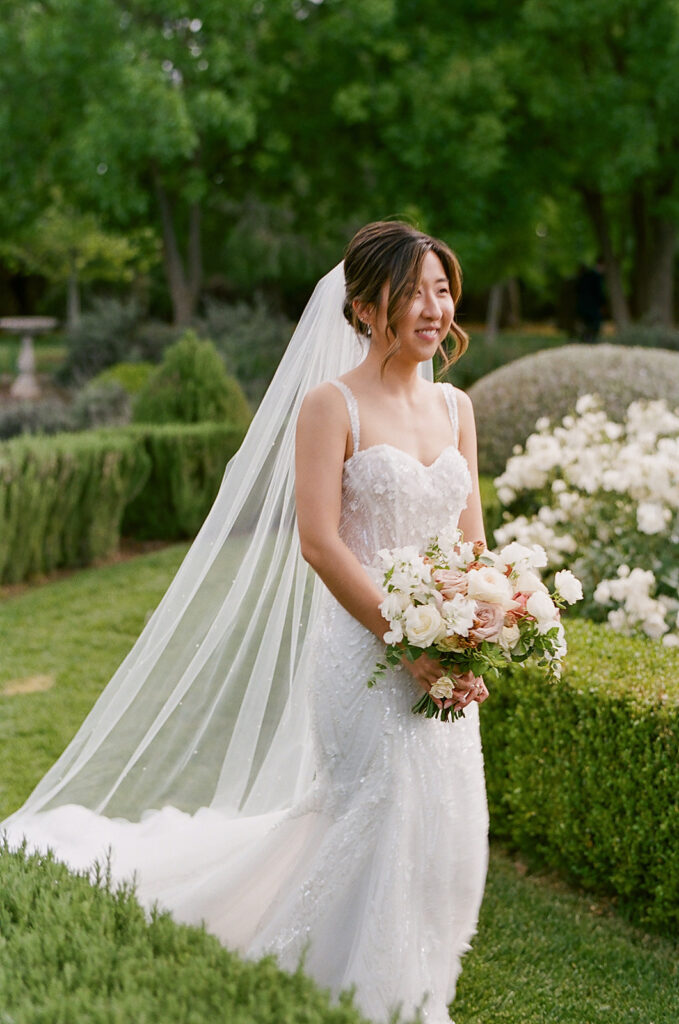 The bride, in a white dress with a delicate veil, holds a bouquet of flowers while standing amidst the enchanting greenery and white blooms of Park Winters Weddings.