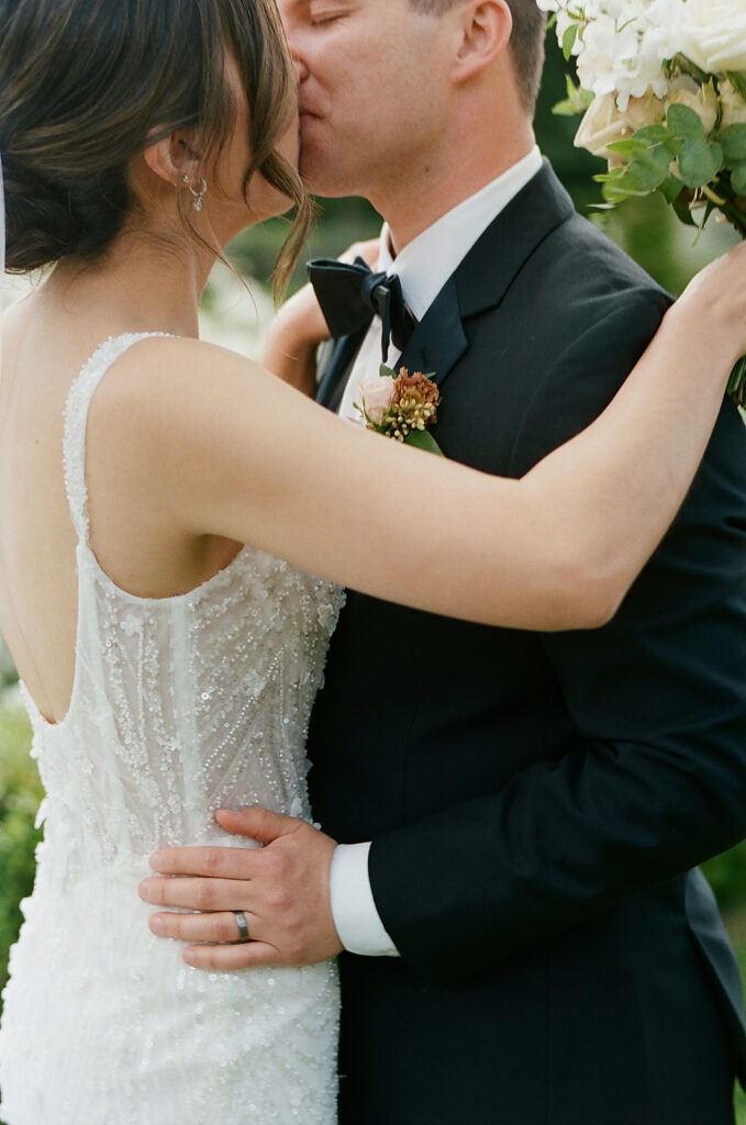 At a Park Winters wedding, a couple in formal attire shares a tender kiss. The woman stuns in a white beaded dress, while the man complements her look with his black suit and floral boutonniere.