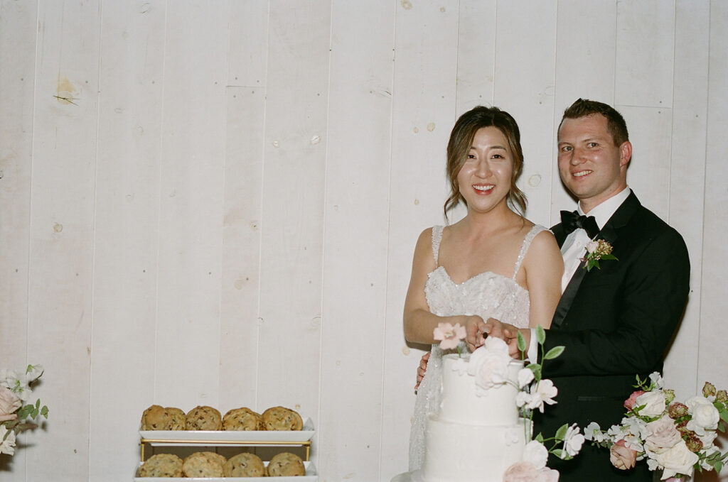 At Park Winters Weddings, the bride and groom beamed as they sliced into their elegant white-tiered cake, a tray of cookies complementing the sweet celebration nearby.