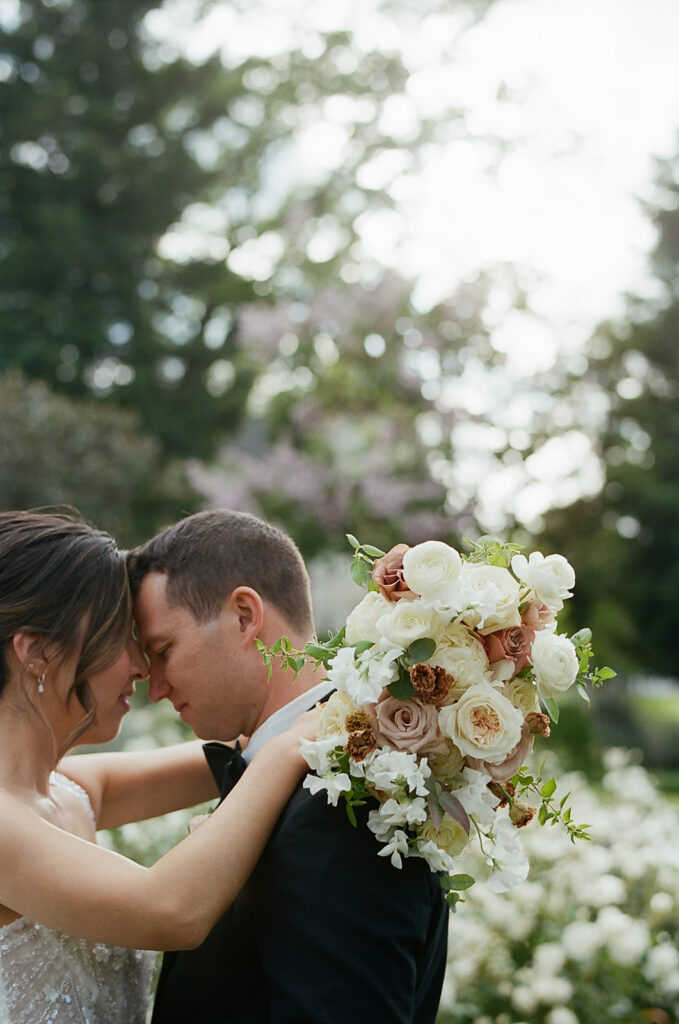 A couple stands closely, touching foreheads at a Park Winters wedding, with a bouquet of white and blush flowers in the foreground, surrounded by greenery and soft lighting.