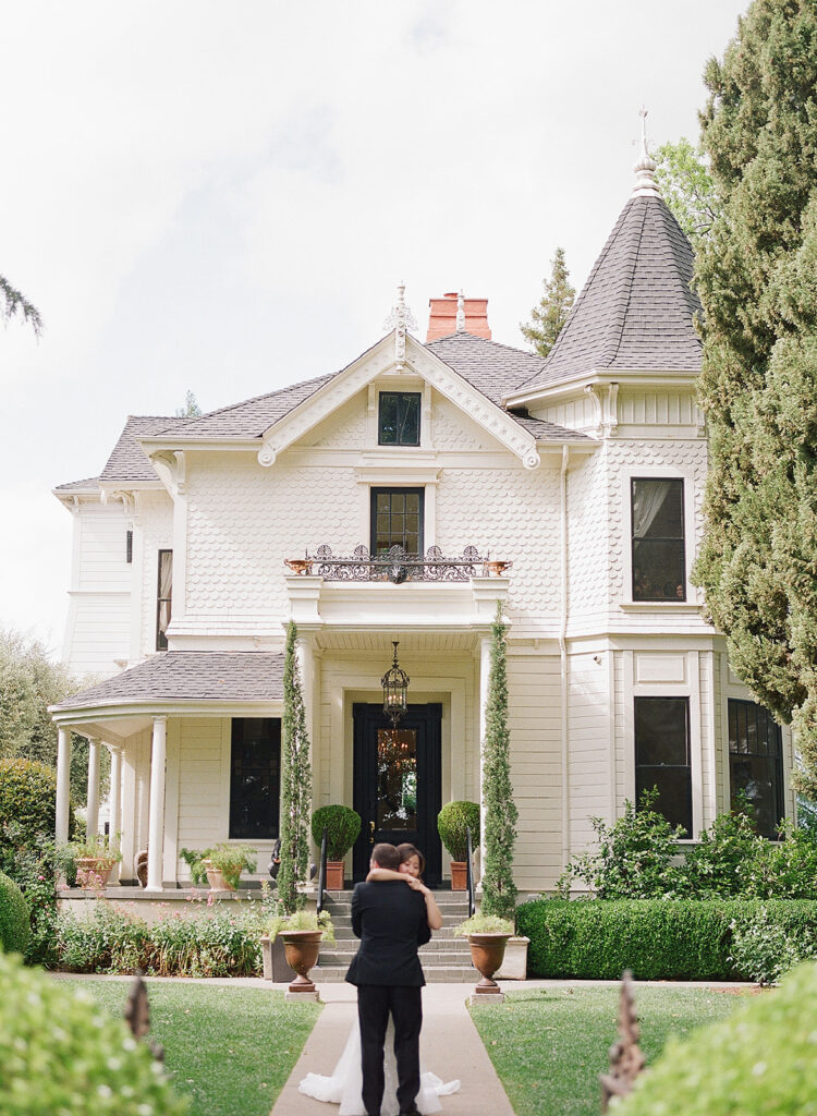 A couple embraces in front of the elegant, two-story Victorian-style house at Park Winters, surrounded by manicured gardens.