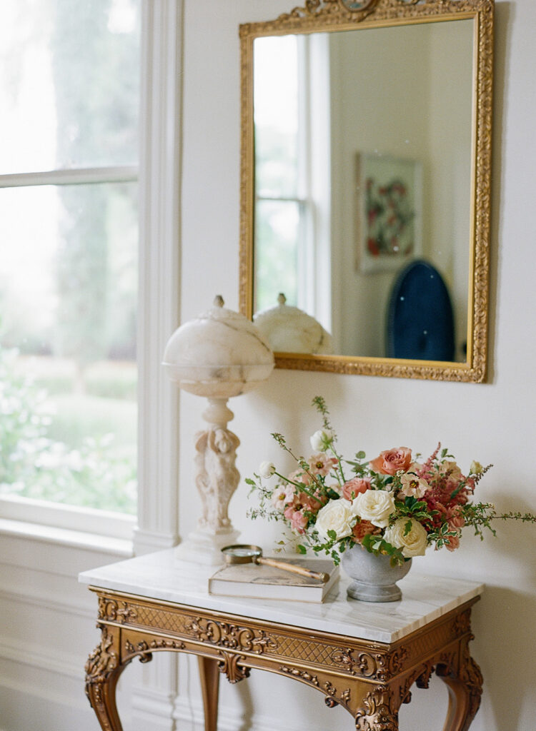 A decorative table with a marble top graces the room, holding a floral arrangement reminiscent of Park Winters Weddings, alongside a small globe lamp and a book. Above it, an ornate gold mirror captures the elegance of such memorable occasions.