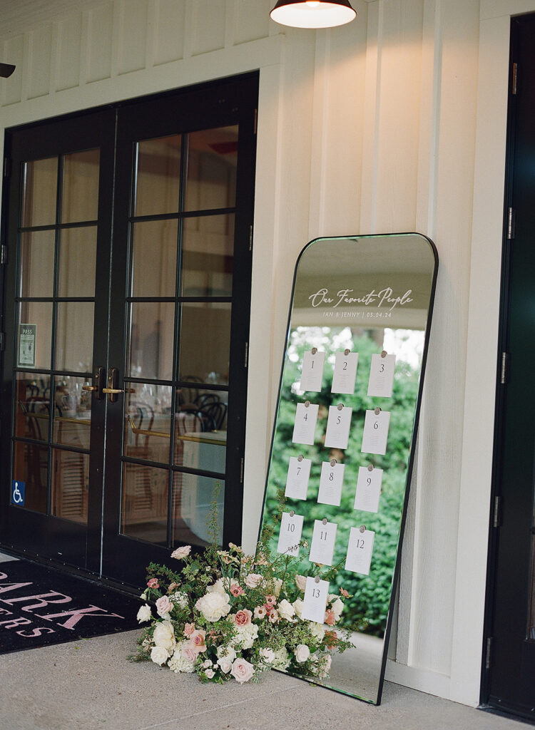 A mirror seating chart with white cards numbered 1 to 13 is beautifully adorned with a flower arrangement at the base, leaning elegantly against the closed glass-paneled doors, capturing the enchanting essence of Park Winters weddings.