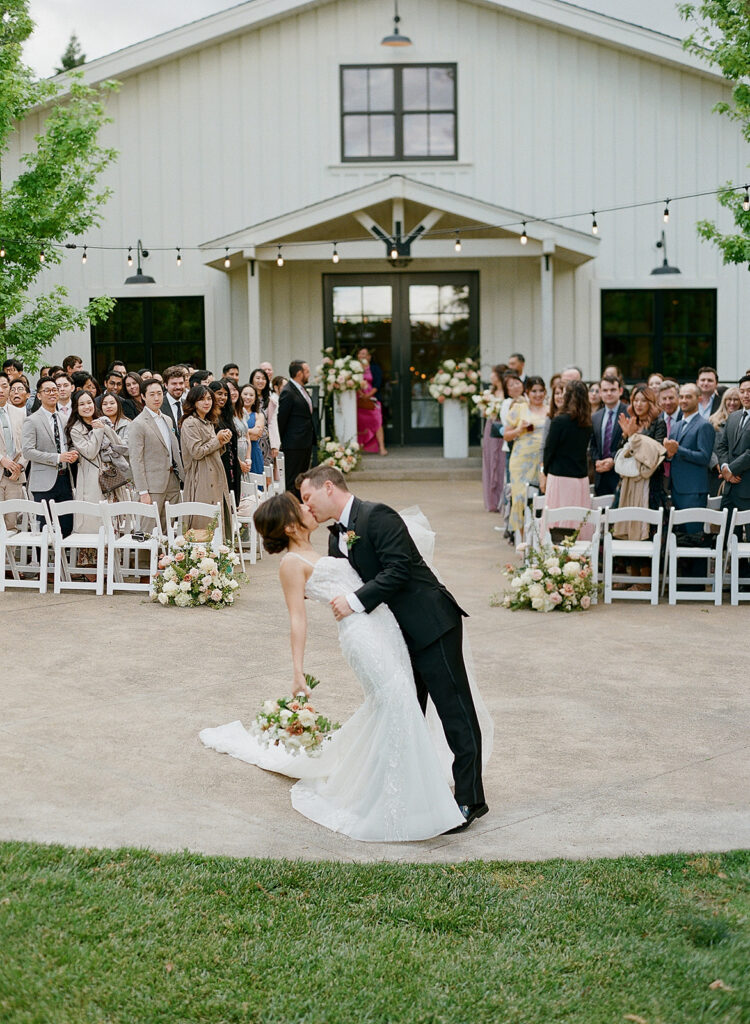 At a Park Winters wedding, a bride and groom share a kiss during their ceremony, surrounded by guests seated in white chairs. The romantic event unfolds outdoors, with the elegant white building providing a stunning backdrop.