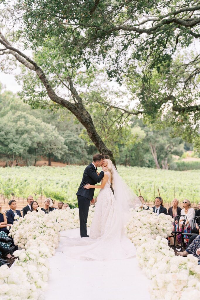 A bride and groom kiss at an outdoor wedding ceremony, surrounded by guests and white flowers, with a vineyard in the background.