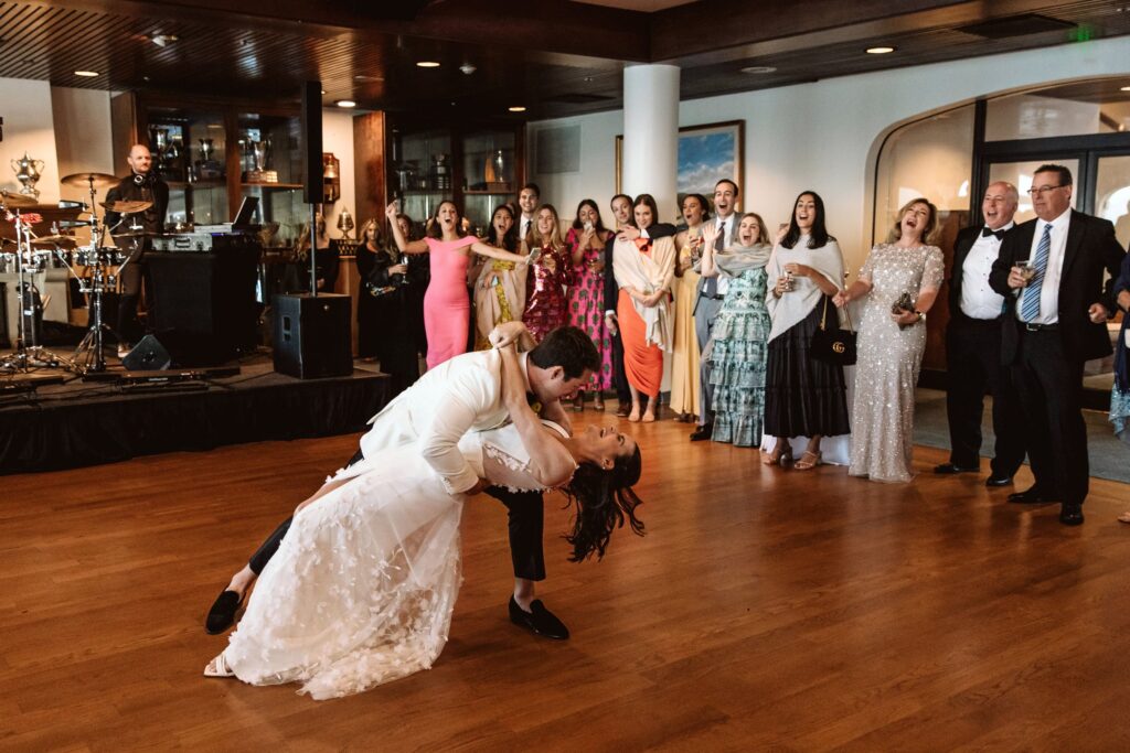 A couple in wedding attire shares a dance while guests watch in a reception hall. A band is set up onstage in the background.