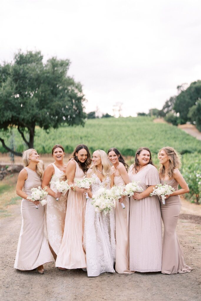 A bride stands with six bridesmaids in pastel dresses, holding bouquets, on a path with greenery and trees in the background.